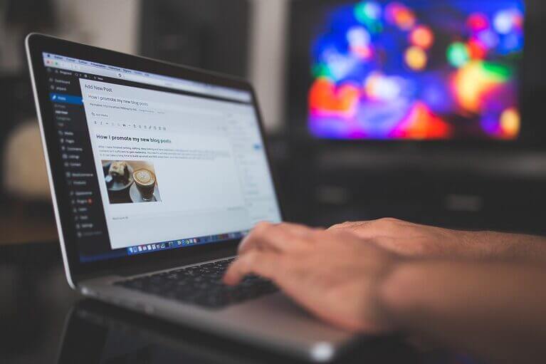 A person multitasking by typing on a laptop while a TV plays in the background.